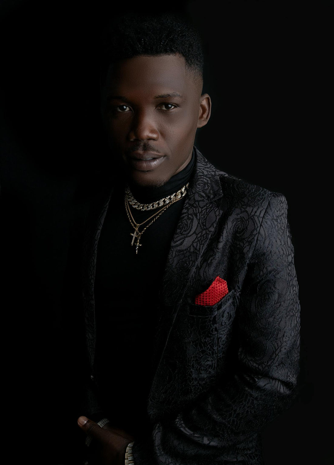 Confident man in a black patterned suit jacket with gold chains and red pocket square against a dark background