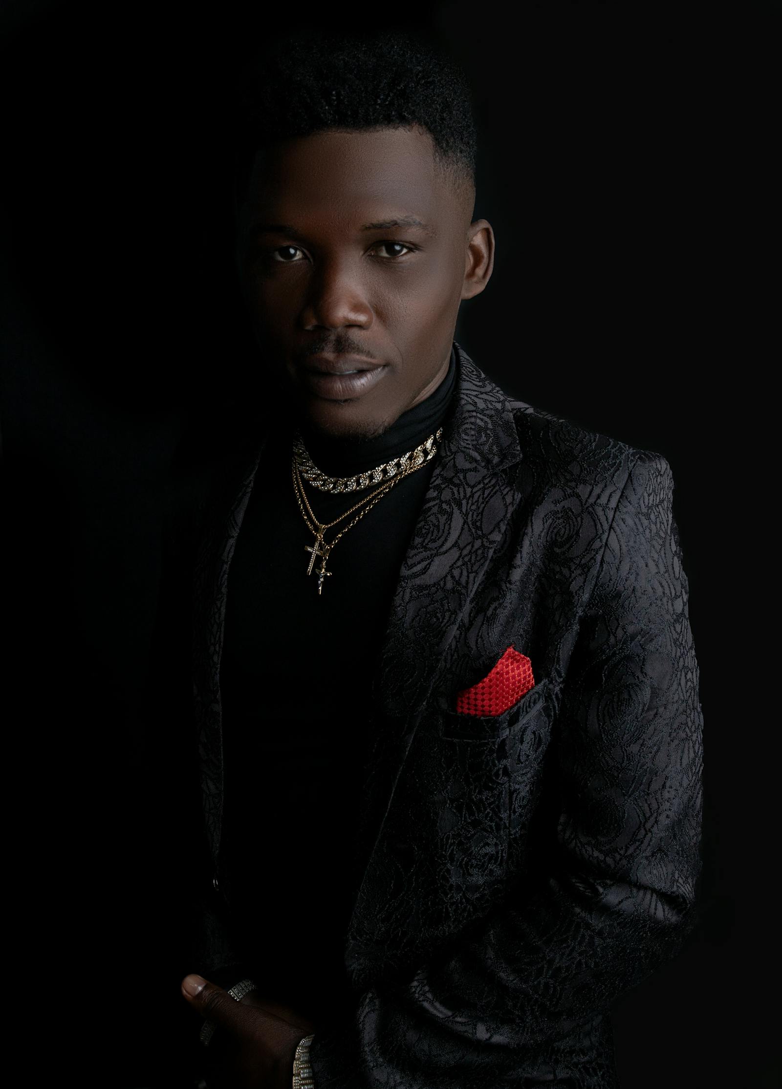 Confident man in a black patterned suit jacket with gold chains and red pocket square against a dark background