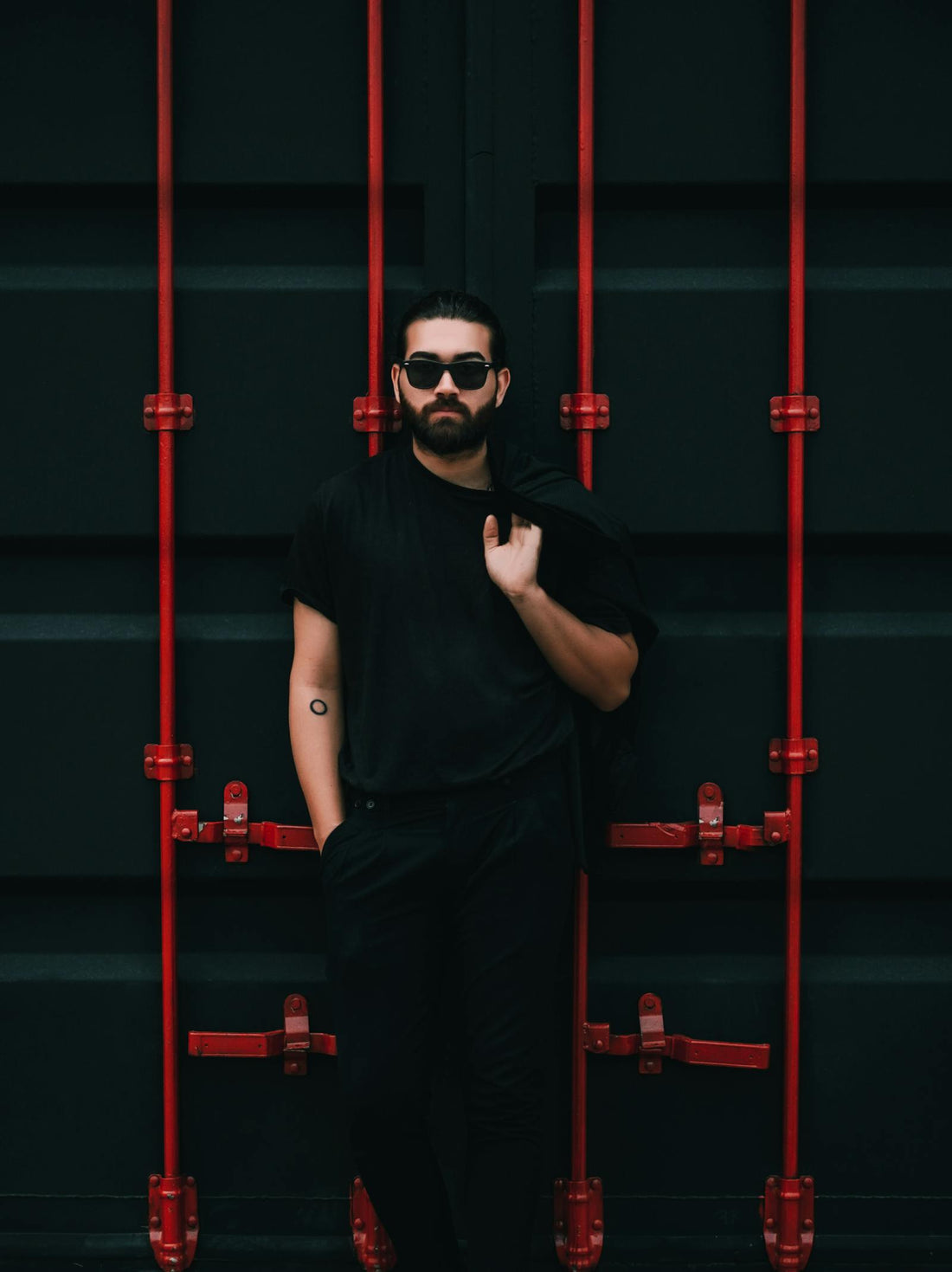 Bearded man in black wearing dark sunglasses, leaning against dark red-accented wall
